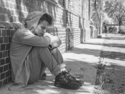 A boy sitting on the street with his hood up.